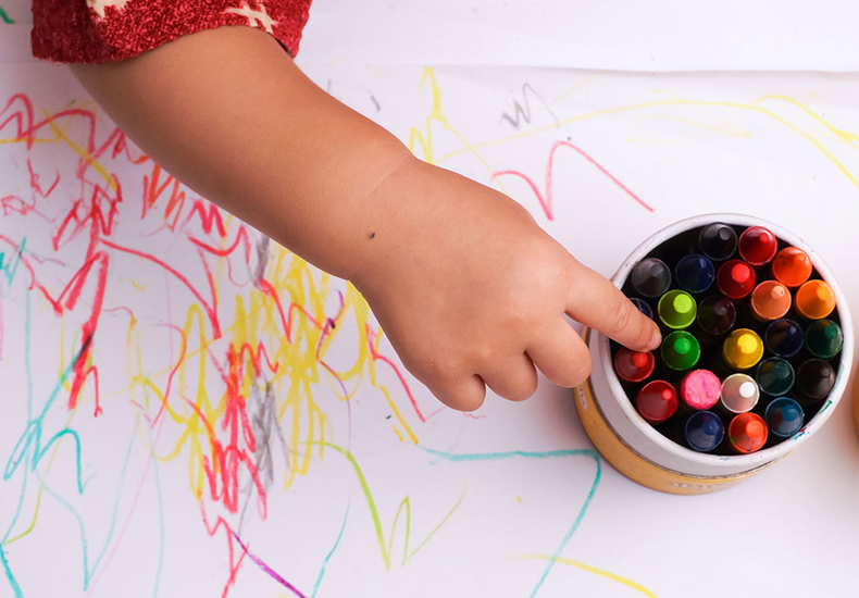 Child's hand reaching over a paper with different coloured scribbles on it for a crayon in a cup of crayons.