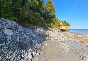 An excavator is moving along the shoreline, placing armourstone along the base of the bluff as it moves from east to west. 